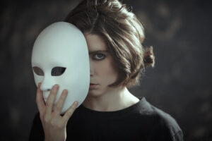 Woman holding white mask partially over face, symbolizing hidden brokenness and emotional pain.